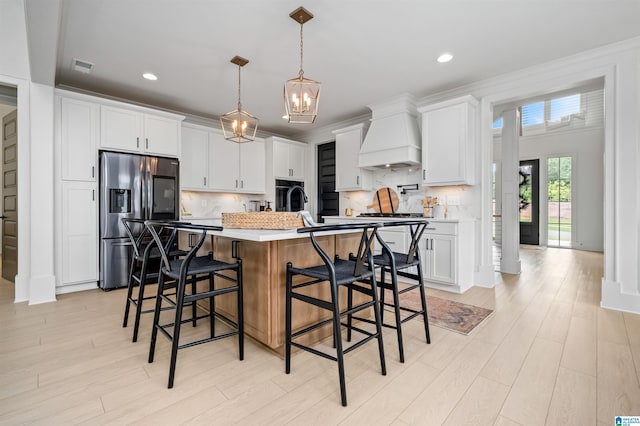 kitchen featuring premium range hood, stainless steel fridge with ice dispenser, hanging light fixtures, a center island with sink, and white cabinets