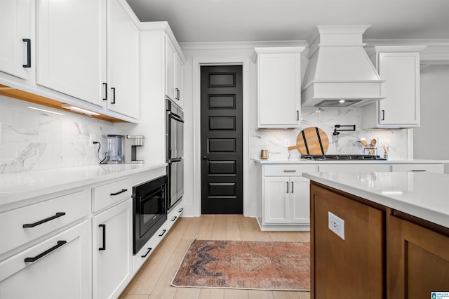 kitchen featuring backsplash, black appliances, custom range hood, white cabinets, and light wood-type flooring