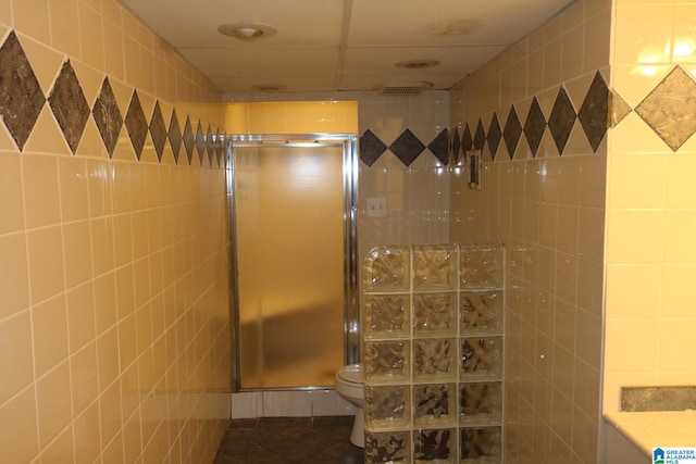 bathroom with a paneled ceiling, toilet, a shower with door, and tile walls