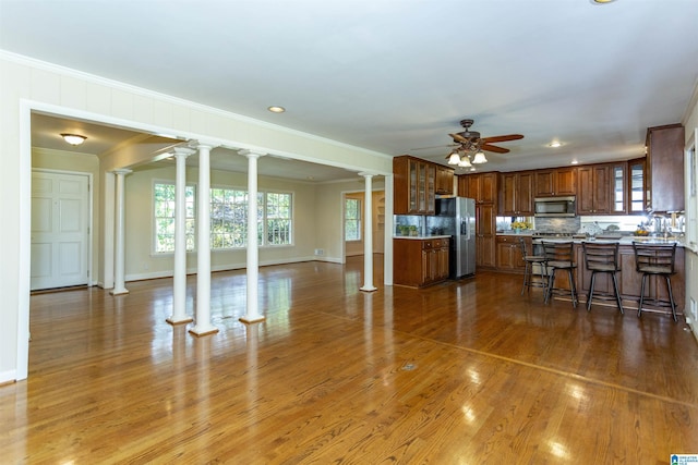 kitchen with glass insert cabinets, appliances with stainless steel finishes, brown cabinets, ornate columns, and a kitchen bar