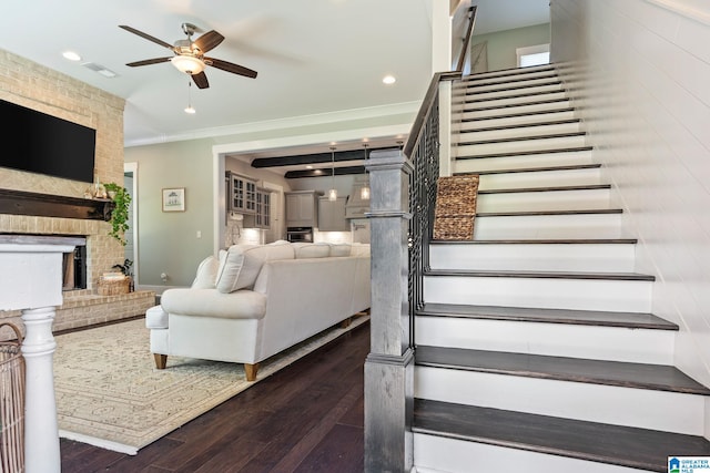 staircase featuring crown molding, dark wood-type flooring, ceiling fan, and a fireplace