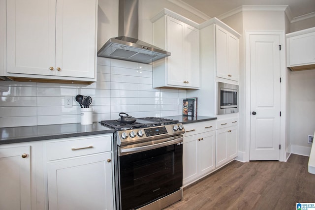 kitchen featuring white cabinets, wall chimney range hood, appliances with stainless steel finishes, dark countertops, and crown molding