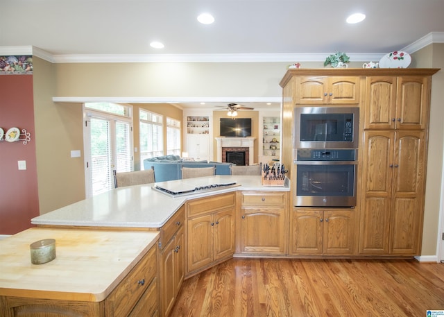 kitchen featuring ceiling fan, stainless steel appliances, a brick fireplace, light hardwood / wood-style flooring, and ornamental molding