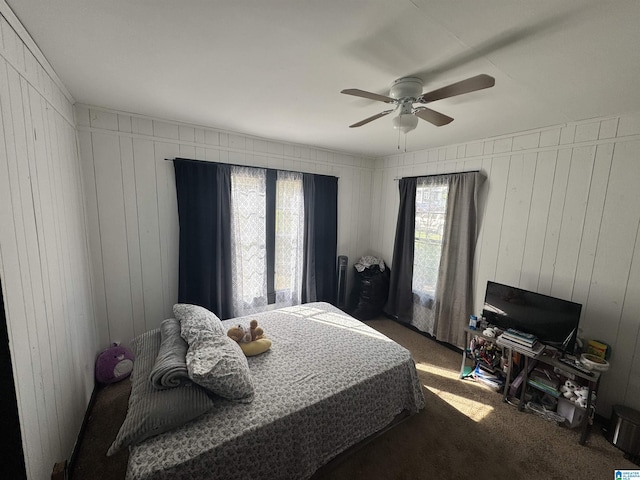 bedroom with ceiling fan, multiple windows, dark carpet, and wood walls