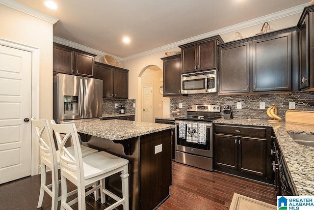 kitchen with stainless steel appliances, a kitchen bar, light stone counters, dark brown cabinets, and backsplash