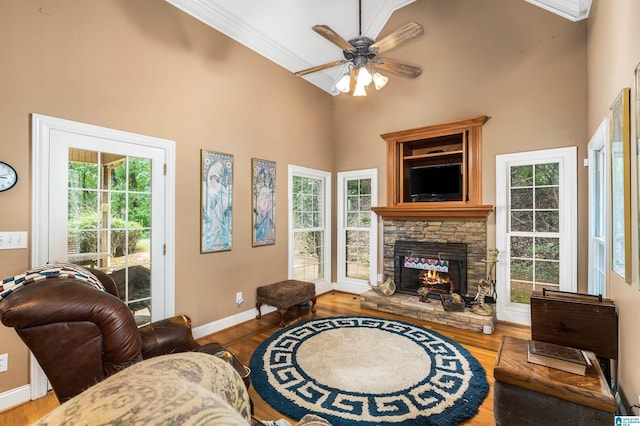 living room featuring a towering ceiling, a ceiling fan, a stone fireplace, wood finished floors, and baseboards