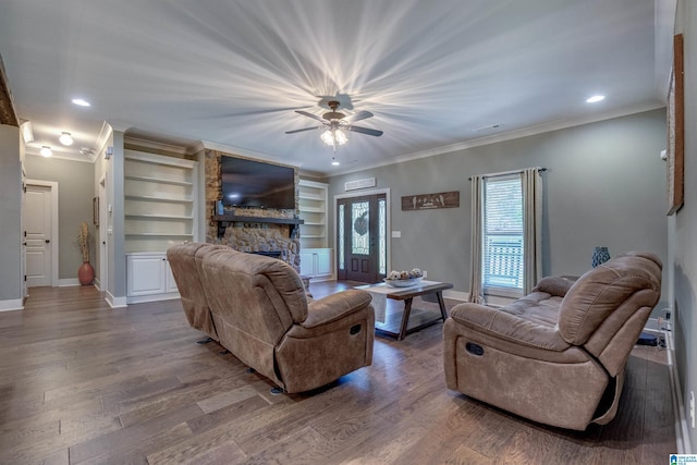 living room with built in features, ceiling fan, wood-type flooring, ornamental molding, and a stone fireplace