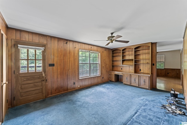 unfurnished living room with ceiling fan, a healthy amount of sunlight, built in desk, and dark colored carpet