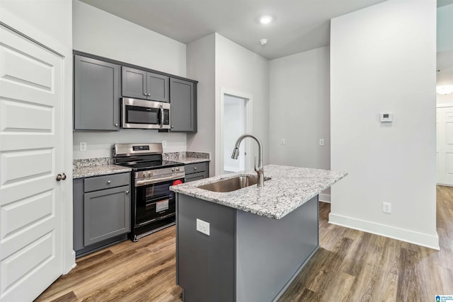 kitchen featuring sink, appliances with stainless steel finishes, gray cabinetry, light stone countertops, and a center island with sink