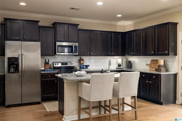 kitchen with light stone counters, light hardwood / wood-style flooring, appliances with stainless steel finishes, a kitchen island with sink, and decorative backsplash