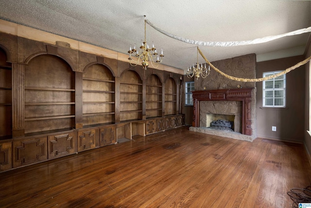 unfurnished living room featuring built in features, a stone fireplace, a textured ceiling, dark hardwood / wood-style flooring, and a notable chandelier