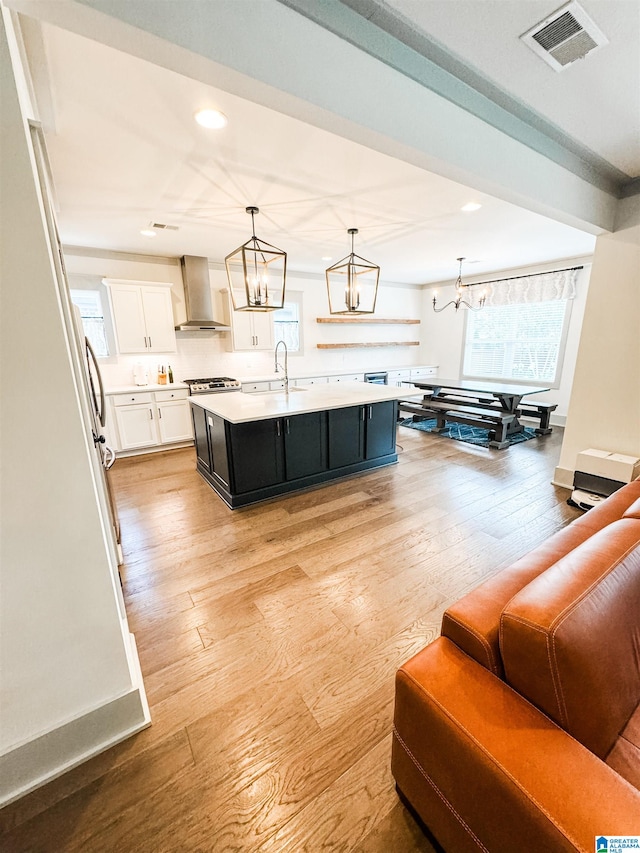 kitchen featuring pendant lighting, white cabinets, a kitchen island with sink, light hardwood / wood-style floors, and wall chimney range hood