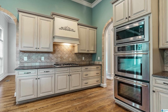 kitchen featuring appliances with stainless steel finishes, tasteful backsplash, dark stone countertops, ornamental molding, and light hardwood / wood-style floors