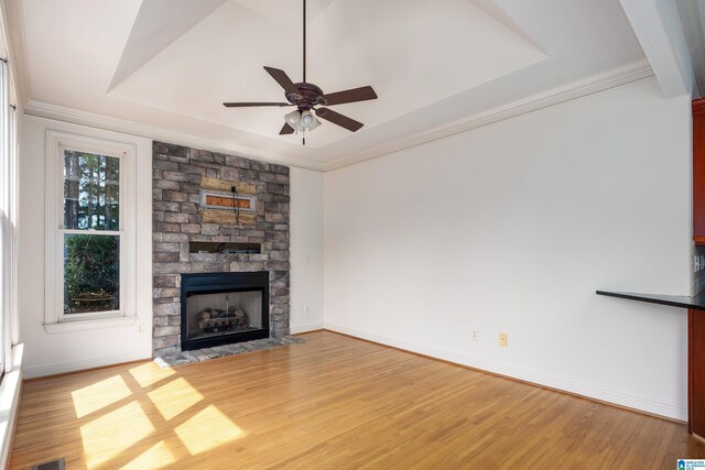 unfurnished living room featuring a stone fireplace, crown molding, light wood-type flooring, ceiling fan, and a raised ceiling