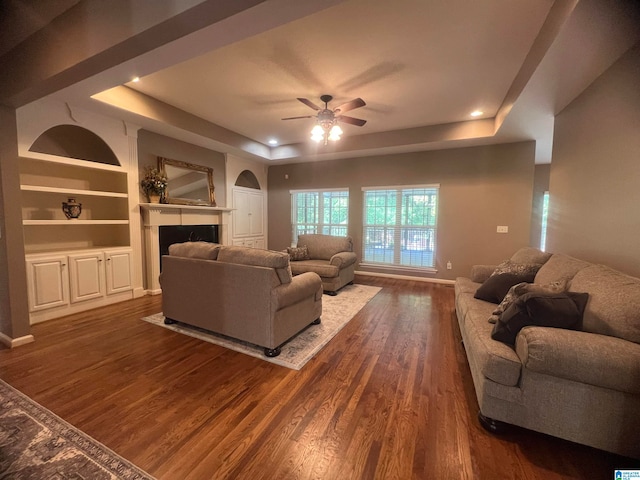 living room with built in features, ceiling fan, wood-type flooring, and a raised ceiling