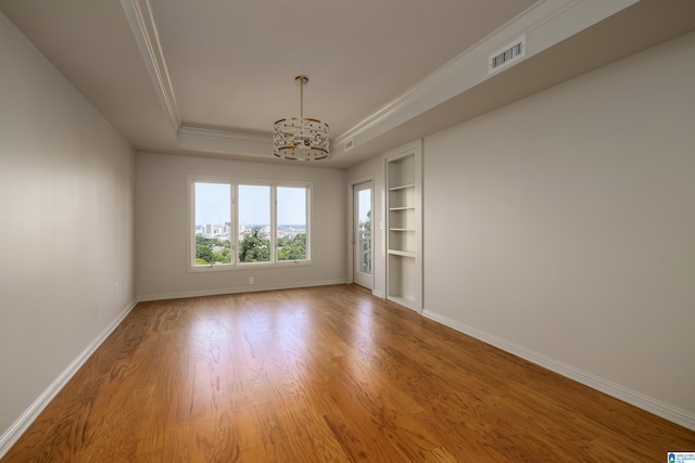 spare room with crown molding, an inviting chandelier, a raised ceiling, built in features, and hardwood / wood-style flooring