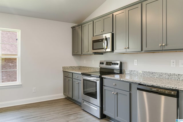 kitchen with stainless steel appliances, light wood-type flooring, vaulted ceiling, gray cabinetry, and light stone counters