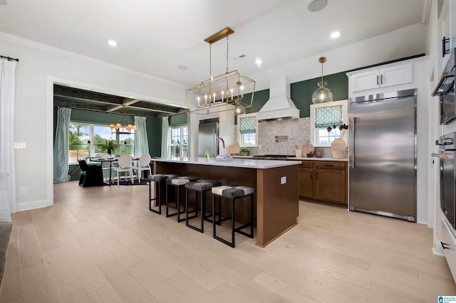 kitchen featuring a kitchen island with sink, appliances with stainless steel finishes, white cabinets, custom exhaust hood, and tasteful backsplash