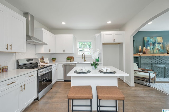 kitchen featuring wall chimney range hood, light wood-type flooring, a brick fireplace, a center island, and stainless steel appliances