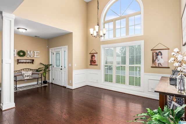 foyer featuring dark wood-type flooring, a chandelier, wainscoting, a towering ceiling, and ornate columns