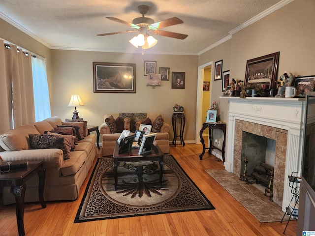 living room featuring ceiling fan, light hardwood / wood-style flooring, a tiled fireplace, a textured ceiling, and crown molding