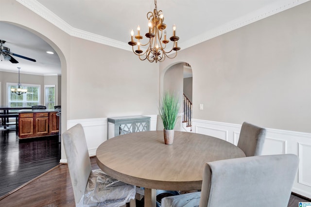 dining space featuring arched walkways, a wainscoted wall, ornamental molding, dark wood-type flooring, and ceiling fan with notable chandelier