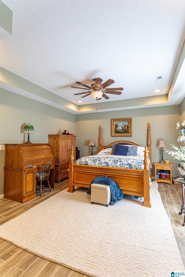 bedroom featuring a textured ceiling, ceiling fan, wood finished floors, visible vents, and a tray ceiling