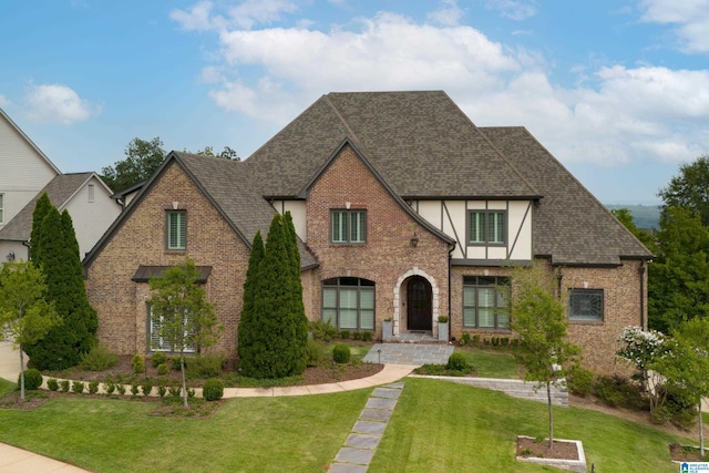 english style home with brick siding, stucco siding, a front yard, and roof with shingles