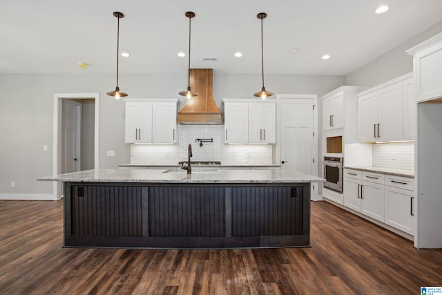 kitchen featuring oven, premium range hood, an island with sink, dark wood-type flooring, and white cabinets