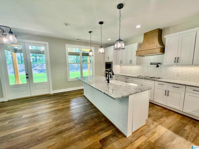 kitchen featuring hardwood / wood-style floors, custom exhaust hood, appliances with stainless steel finishes, a center island with sink, and white cabinets