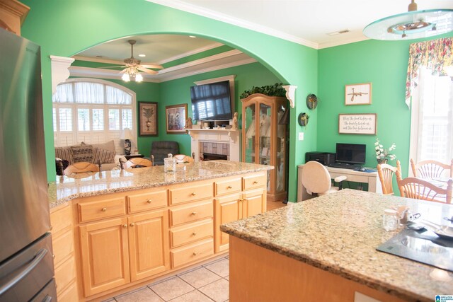 kitchen with a tile fireplace, crown molding, light stone countertops, stainless steel refrigerator, and ceiling fan