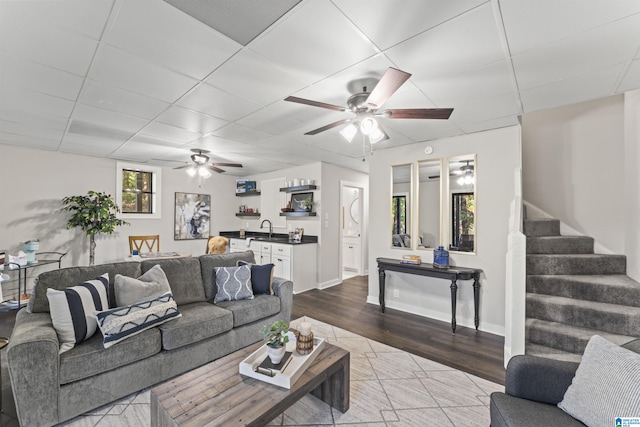 living area featuring stairs, ceiling fan, wood finished floors, and a paneled ceiling