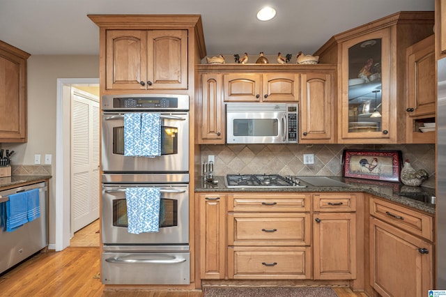 kitchen with light wood-type flooring, appliances with stainless steel finishes, backsplash, and dark stone counters