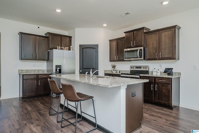 kitchen with light stone countertops, dark hardwood / wood-style flooring, appliances with stainless steel finishes, an island with sink, and sink