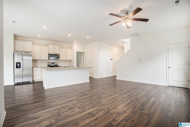 kitchen with light stone countertops, stainless steel appliances, dark wood-type flooring, ceiling fan, and a center island with sink