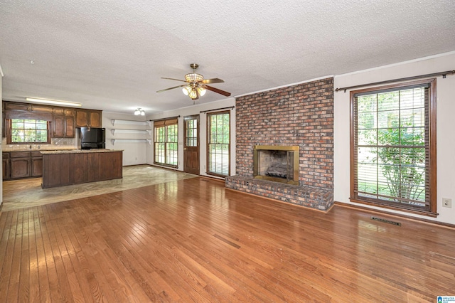 unfurnished living room with light wood-type flooring, a textured ceiling, a brick fireplace, and ceiling fan