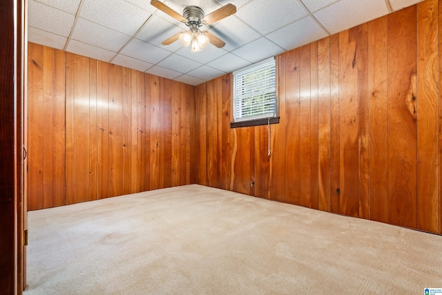 carpeted empty room with ceiling fan, a paneled ceiling, and wood walls