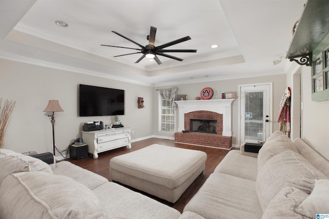 living room featuring dark wood-type flooring, a brick fireplace, a raised ceiling, ornamental molding, and ceiling fan