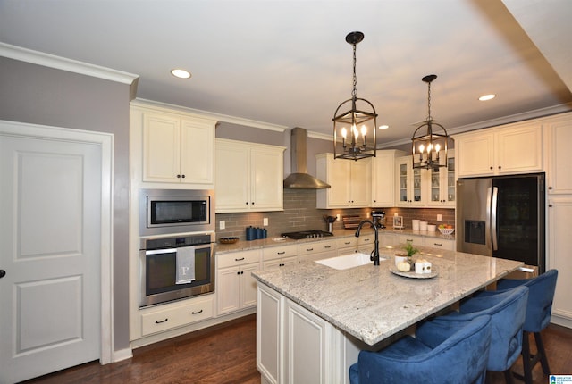 kitchen with stainless steel appliances, a notable chandelier, sink, wall chimney exhaust hood, and a kitchen island with sink