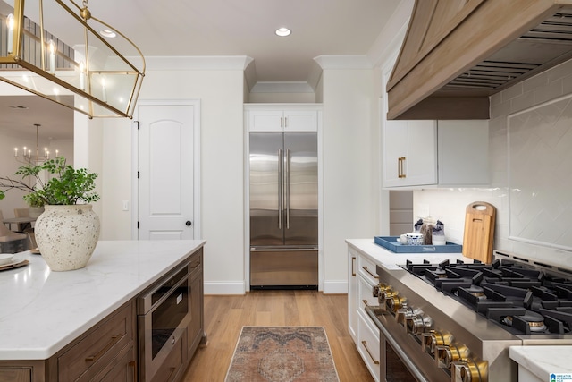 kitchen with premium range hood, built in appliances, light stone counters, a notable chandelier, and white cabinetry