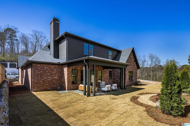 back of property with a patio, brick siding, roof with shingles, and a chimney