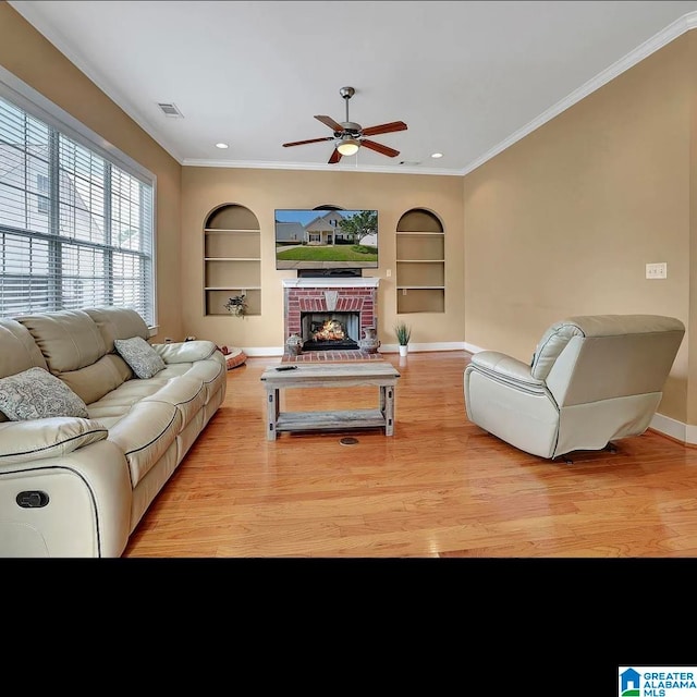 living room featuring built in shelves, ceiling fan, a brick fireplace, light hardwood / wood-style flooring, and crown molding