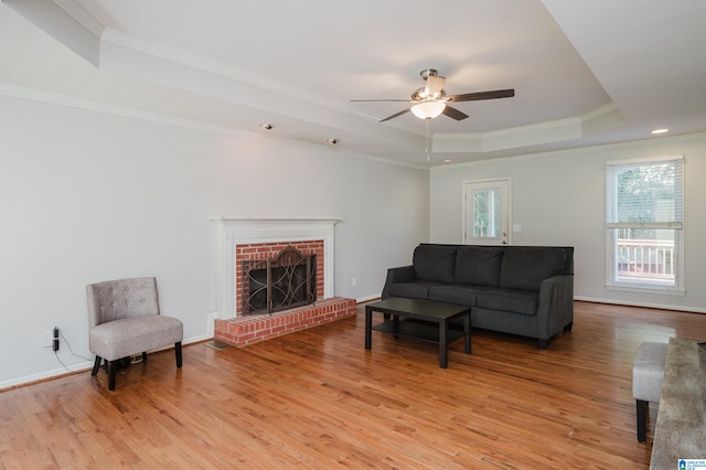 living room with ceiling fan, a raised ceiling, a brick fireplace, crown molding, and light hardwood / wood-style floors