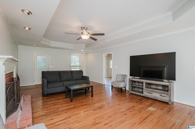 living room featuring ceiling fan, a raised ceiling, ornamental molding, a brick fireplace, and light hardwood / wood-style flooring