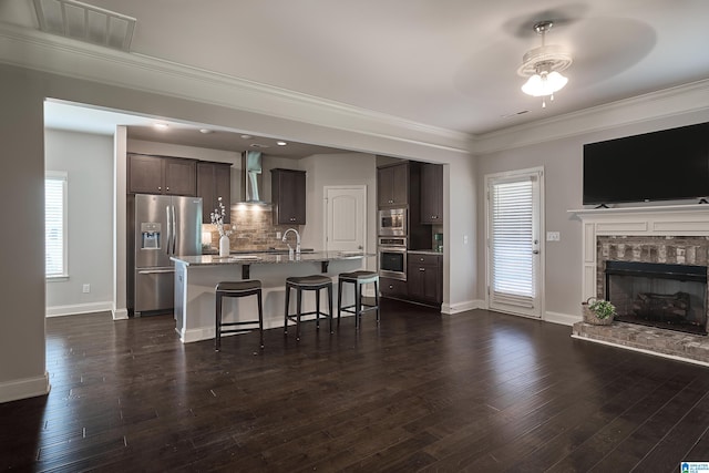 kitchen with ceiling fan, a kitchen island with sink, wall chimney range hood, stainless steel appliances, and a fireplace