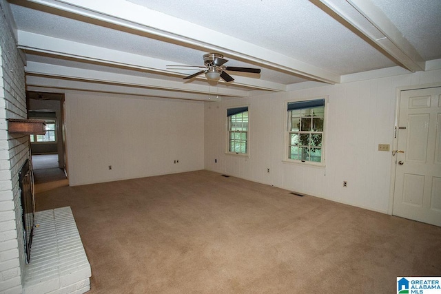 carpeted empty room with a brick fireplace, ceiling fan, beamed ceiling, and a textured ceiling