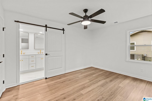 unfurnished bedroom featuring ceiling fan, sink, a barn door, light wood-type flooring, and ensuite bathroom