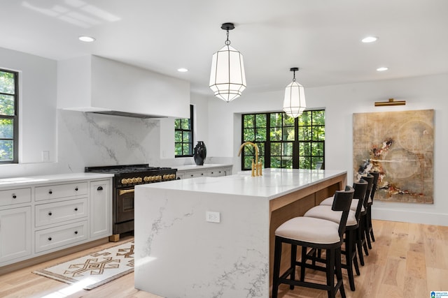 kitchen with white cabinets, custom exhaust hood, a center island with sink, stainless steel range, and light wood-type flooring