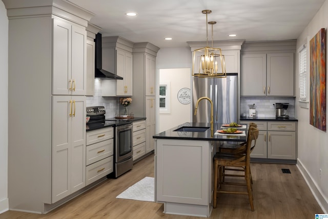 kitchen featuring wall chimney range hood, light hardwood / wood-style flooring, a center island with sink, hanging light fixtures, and appliances with stainless steel finishes