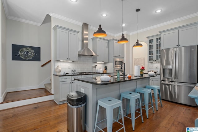 kitchen with an island with sink, stainless steel appliances, wall chimney exhaust hood, and dark wood-type flooring